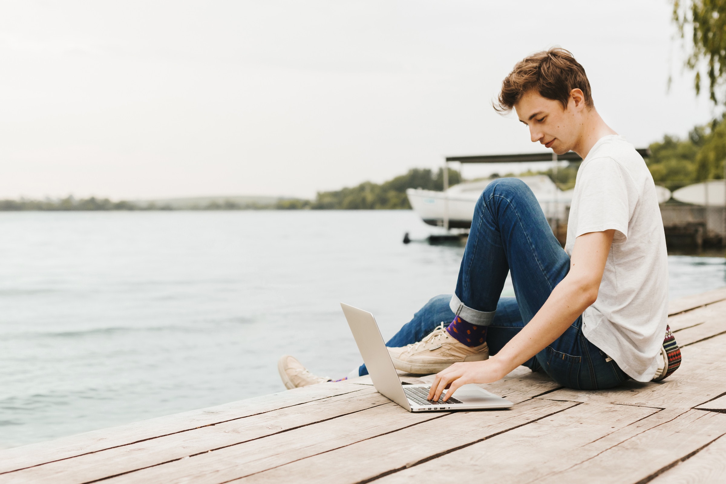 Person sitter på en brygga vid vattnet och beställer på sin laptop, med en båt i bakgrunden