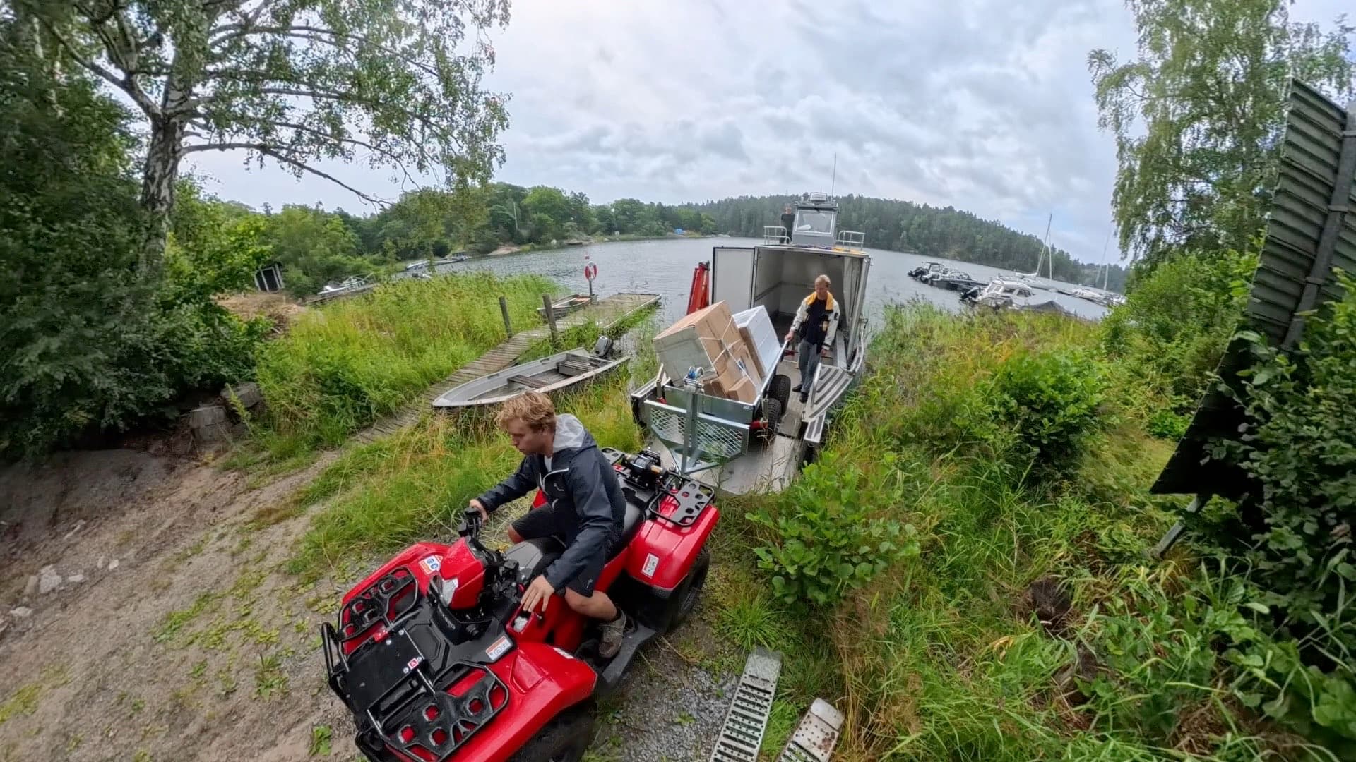 Fyrhjuling med släp lastat med kartonger på väg upp från båten vid en gräsbevuxen strand